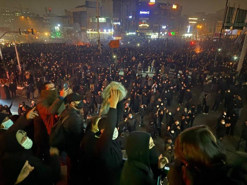 A large crowd of protesters fills a city intersection at night, with hundreds of people dressed mostly in dark clothing. Many wear face masks and hold up phones, while small fires or flares glow in the distance. Bright shop signs and traffic lights illuminate the scene